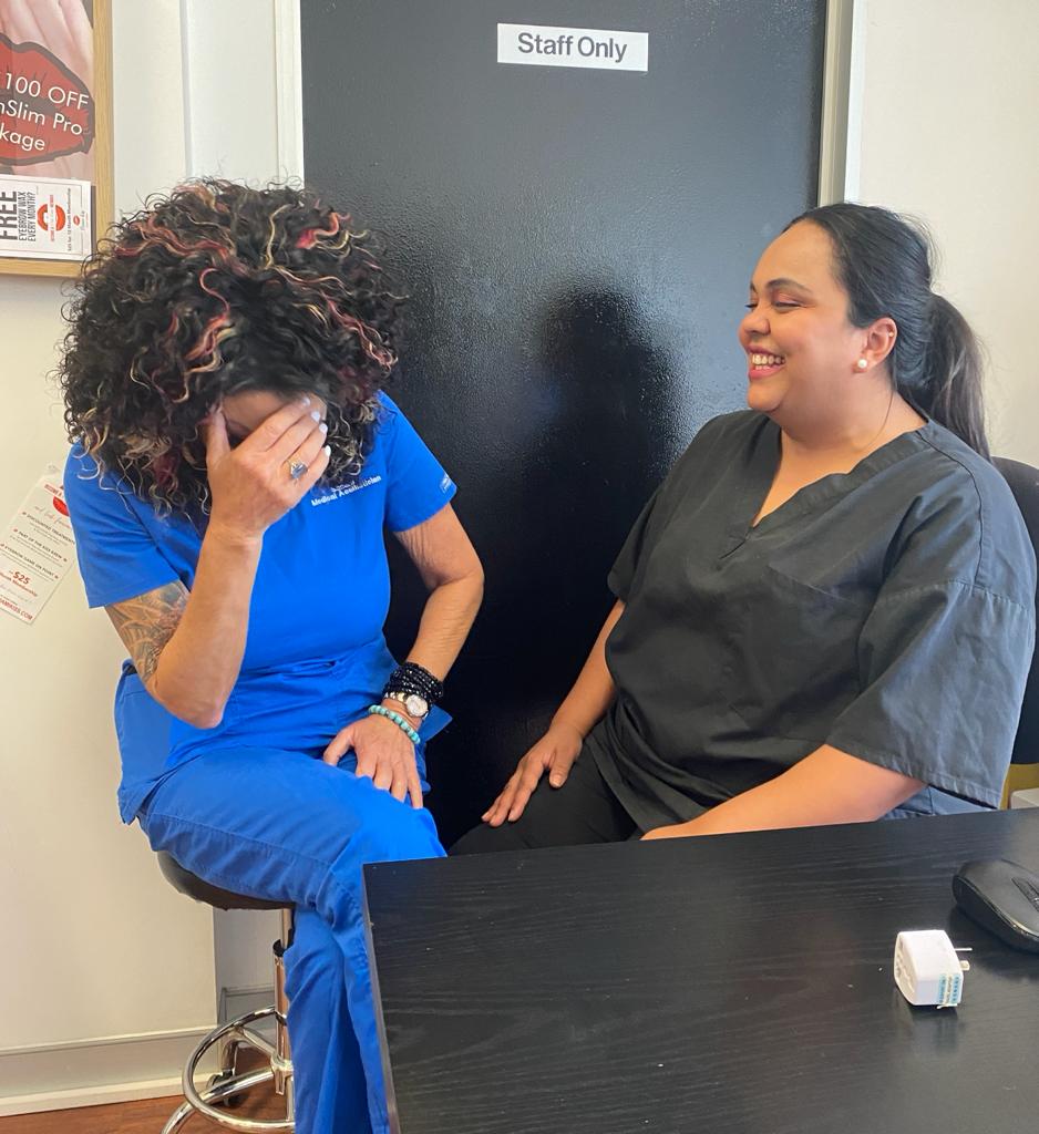 image of Sandra, Founder of Miami Kiss a woman wearing blue medical scrubs and Anushka Owner of Miami Kiss and registered nurse wearing black medical scrubs sitting at desk laughing