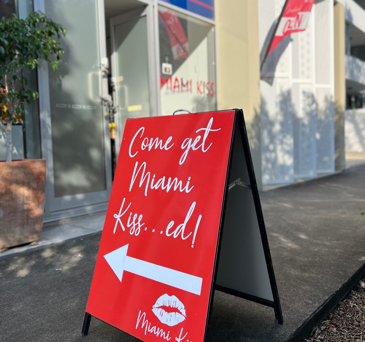 Exterior photo of Miami Kiss clinic with a-frame in foreground reading 'Come get Miami Kissed' and shopfront in background